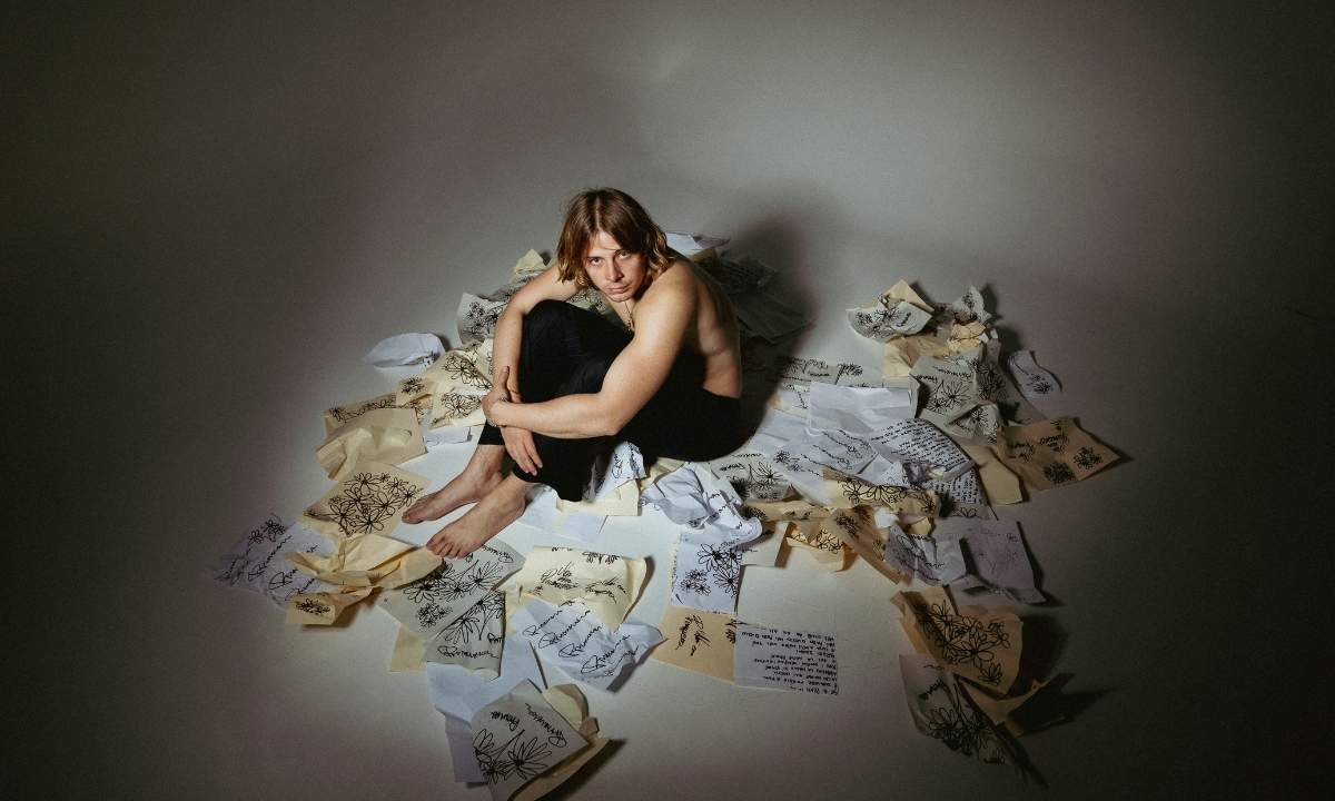 FARES seduto a terra circondato da fogli con fiori e testi scritti a mano, scatto promozionale del singolo "Primavera"