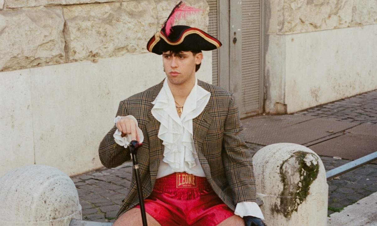 Primogenito seduto su un marciapiede con cappello a tricorno, giacca a quadri e pantaloncini da boxe rossi