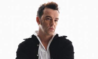 Salmo in posa su sfondo bianco, indossa una camicia bianca sbottonata e una giacca nera aperta, con i tatuaggi sulle mani in evidenza.