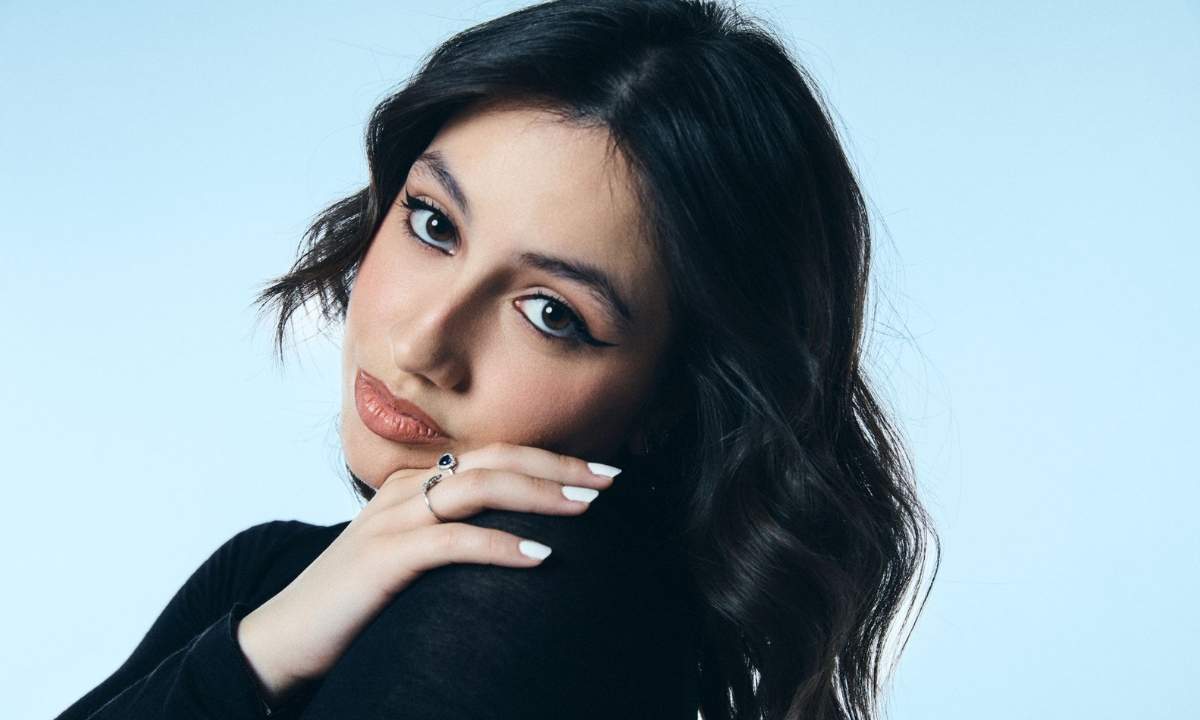 Angie posa con la mano sul viso, capelli lunghi castani e maglia nera, su sfondo azzurro chiaro