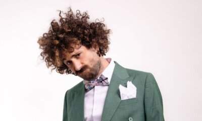 Gio Evan in giacca verde, papillon e pochette bianca, capelli ricci, sguardo verso il basso su sfondo bianco