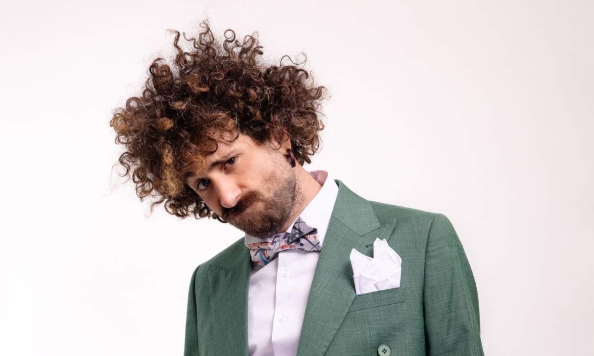 Gio Evan in giacca verde, papillon e pochette bianca, capelli ricci, sguardo verso il basso su sfondo bianco