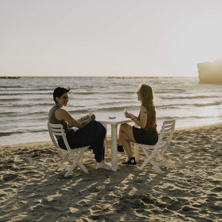 Cover del singolo Quando poi ci lasceremo di Giordana Angi: due figure femminili a un tavolo su una spiaggia al tramonto. 