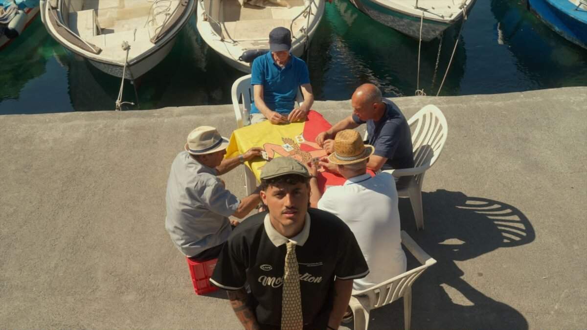 Saitta in primo piano davanti a un tavolo di anziani che giocano a carte su una tovaglia con la Trinacria siciliana, in un porto