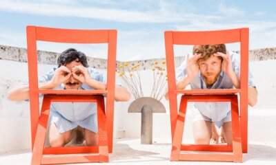 Massimo Di Lena e Lucio Aquilina dei Nu Genea giocano a fare i binocoli con le mani dietro due sedie rosse su una terrazza mediterranea