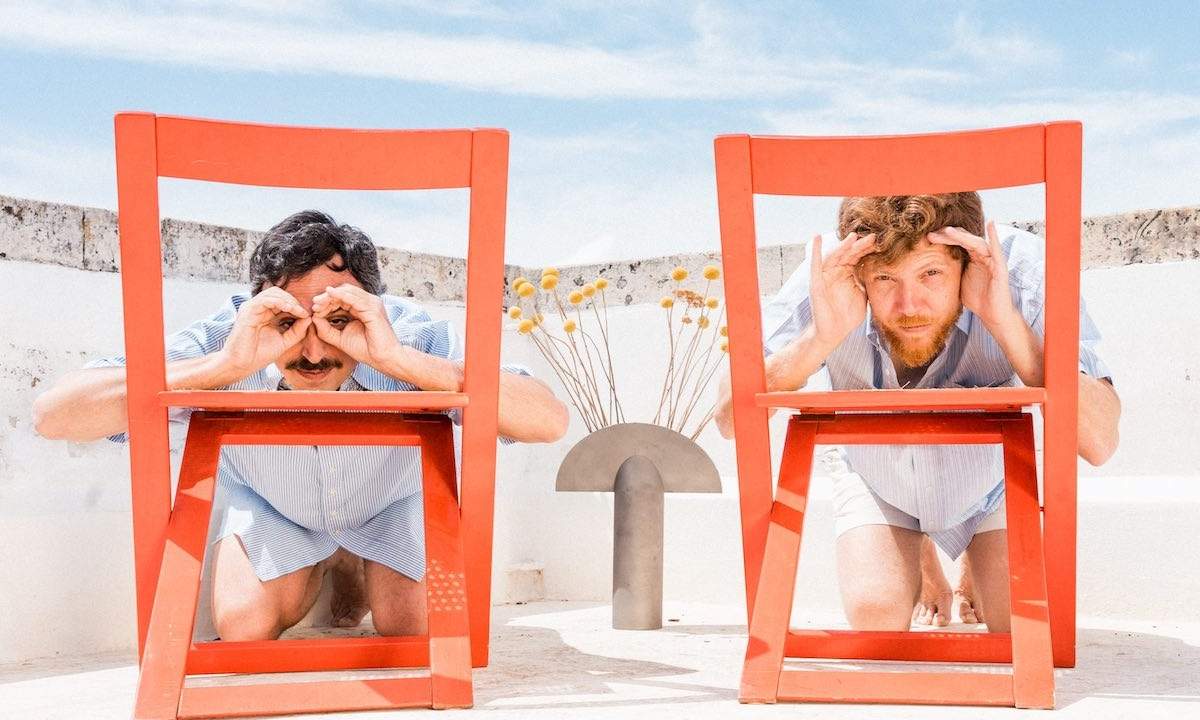 Massimo Di Lena e Lucio Aquilina dei Nu Genea giocano a fare i binocoli con le mani dietro due sedie rosse su una terrazza mediterranea