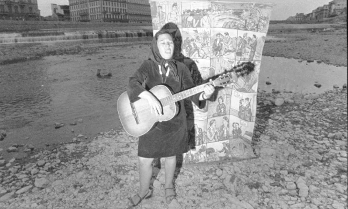 Rosa Balistreri canta con la chitarra sul greto dell'Arno a Firenze, accanto ai cartelloni dei cantastorie