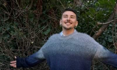 Cricca sorride a braccia aperte davanti a una siepe, con un maglione di lana grigio e blu