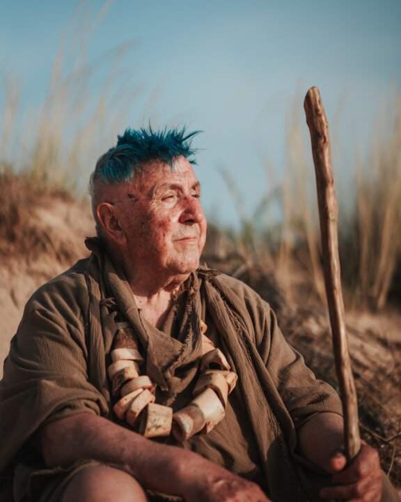 Uomo anziano con mohawk blu, bastone di legno e collana tribale su una duna: copertina di Abbabula, il singolo di DJ BIGZI e J€SS