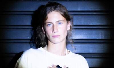 Eva Bloo in un ritratto ravvicinato, capelli mossi e maglietta bianca, davanti a una saracinesca blu