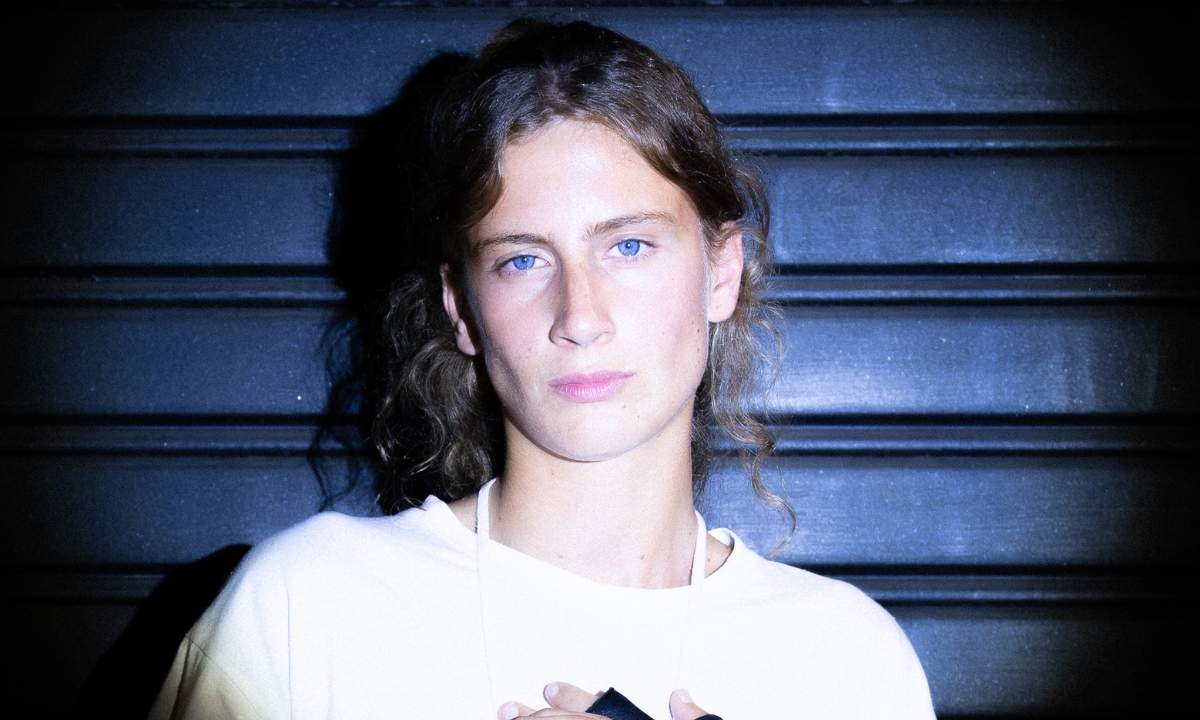 Eva Bloo in un ritratto ravvicinato, capelli mossi e maglietta bianca, davanti a una saracinesca blu