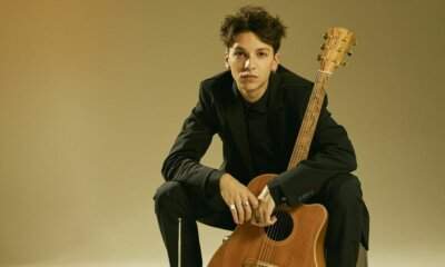 Luigi Strangis seduto con la chitarra acustica, in completo nero su fondo beige