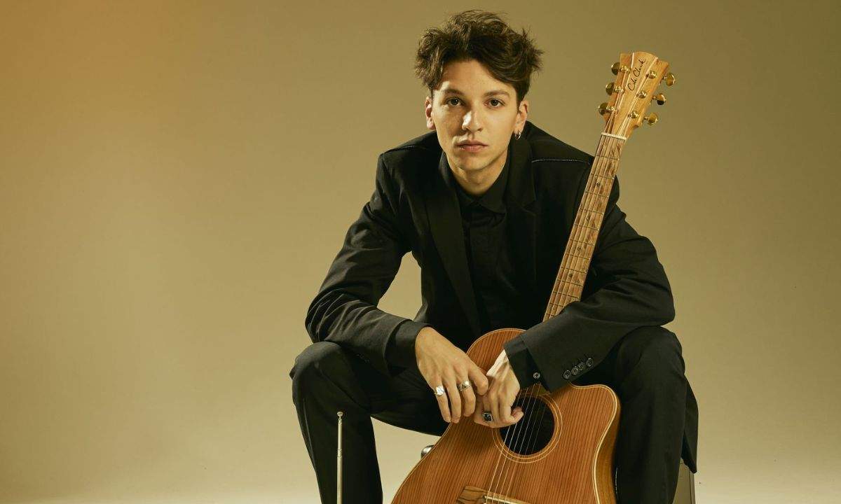 Luigi Strangis seduto con la chitarra acustica, in completo nero su fondo beige