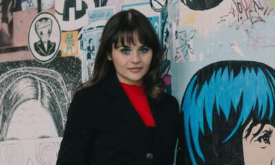Martina Attili in giacca nera e maglia rossa, davanti a un muro coperto di poster e adesivi street art