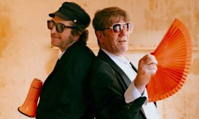 Vinicio Capossela e Pierpaolo Capovilla di spalle uno all'altro con megafono e ventaglio arancioni