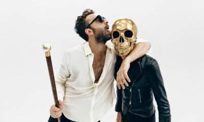 Cesare Cremonini con occhiali scuri e bastone da passeggio accanto a una figura con un teschio d'oro al posto della testa