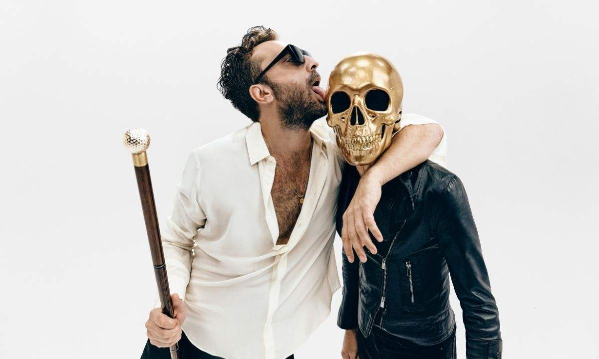 Cesare Cremonini con occhiali scuri e bastone da passeggio accanto a una figura con un teschio d'oro al posto della testa