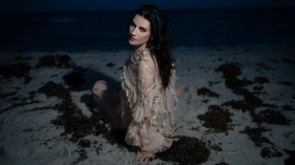 Laura Pausini seduta sulla sabbia di una spiaggia di notte, con un abito di pizzo color cipria e i capelli scuri sciolti