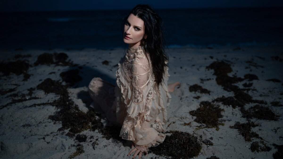 Laura Pausini seduta sulla sabbia di una spiaggia di notte, con un abito di pizzo color cipria e i capelli scuri sciolti
