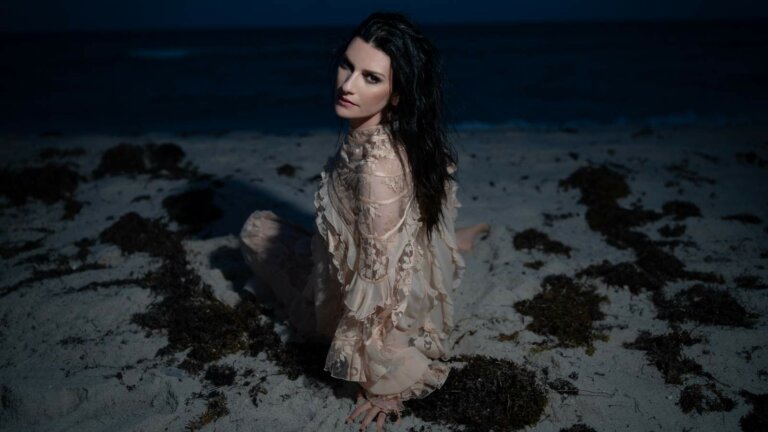 Laura Pausini seduta sulla sabbia di una spiaggia di notte, con un abito di pizzo color cipria e i capelli scuri sciolti