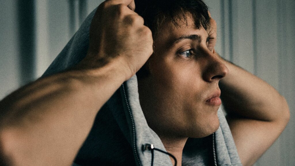 sangiovanni nella foto promozionale dell'album TUTTO BENE GRAZIE, di profilo con le mani sul cappuccio della felpa grigia