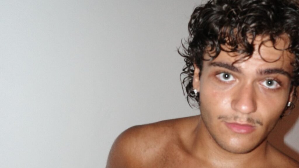 Andrea Heros, il cantautore napoletano entrato ad Amici 26, in un primo piano a torso nudo davanti a un muro bianco