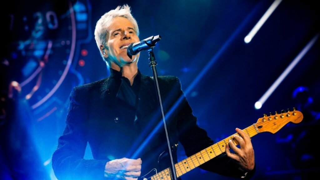Claudio Baglioni durante un'esibizione dal vivo mentre canta al microfono e suona la chitarra
