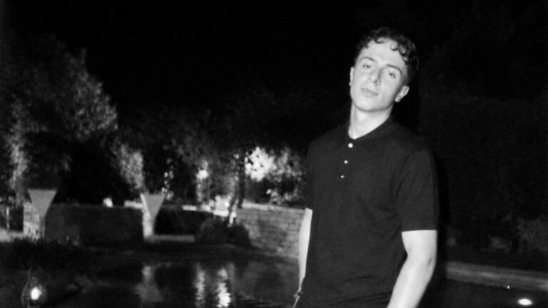 Erness, cantante di Amici 26, in bianco e nero di notte con una polo scura vicino a uno specchio d'acqua