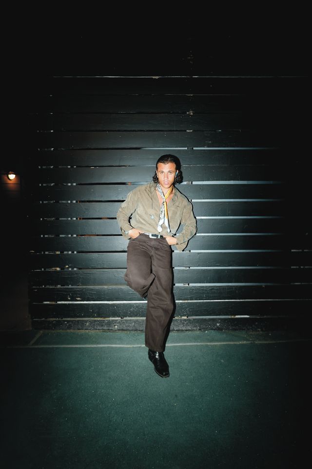 Samurai Jay in un ritratto notturno con il flash, in giacca verde e pantaloni marroni, appoggiato a una parete di assi scure