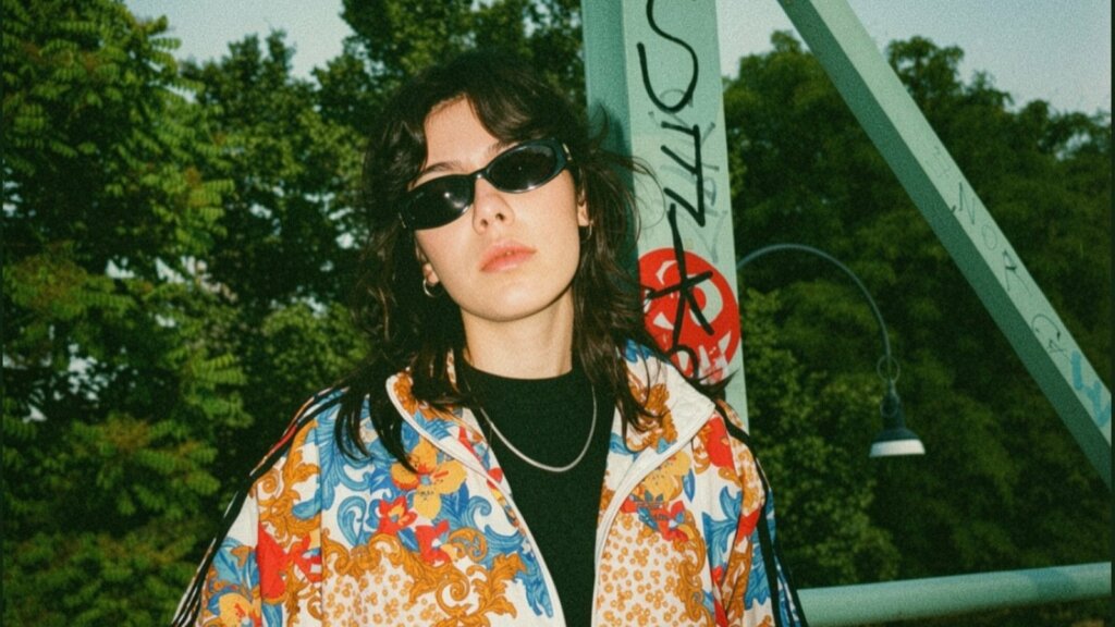 Tìaso, la cantautrice di Caltanissetta entrata ad Amici 26, con occhiali scuri e giacca a fiori davanti a un ponte verde