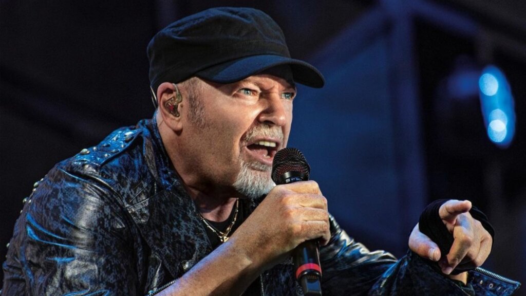 Vasco Rossi in concerto con berretto nero e giubbotto di pelle borchiato, canta nel microfono e indica verso il pubblico