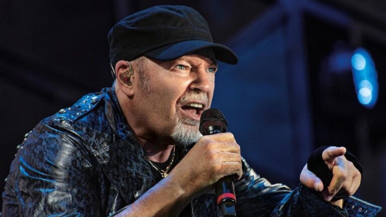 Vasco Rossi in concerto con berretto nero e giubbotto di pelle borchiato, canta nel microfono e indica verso il pubblico